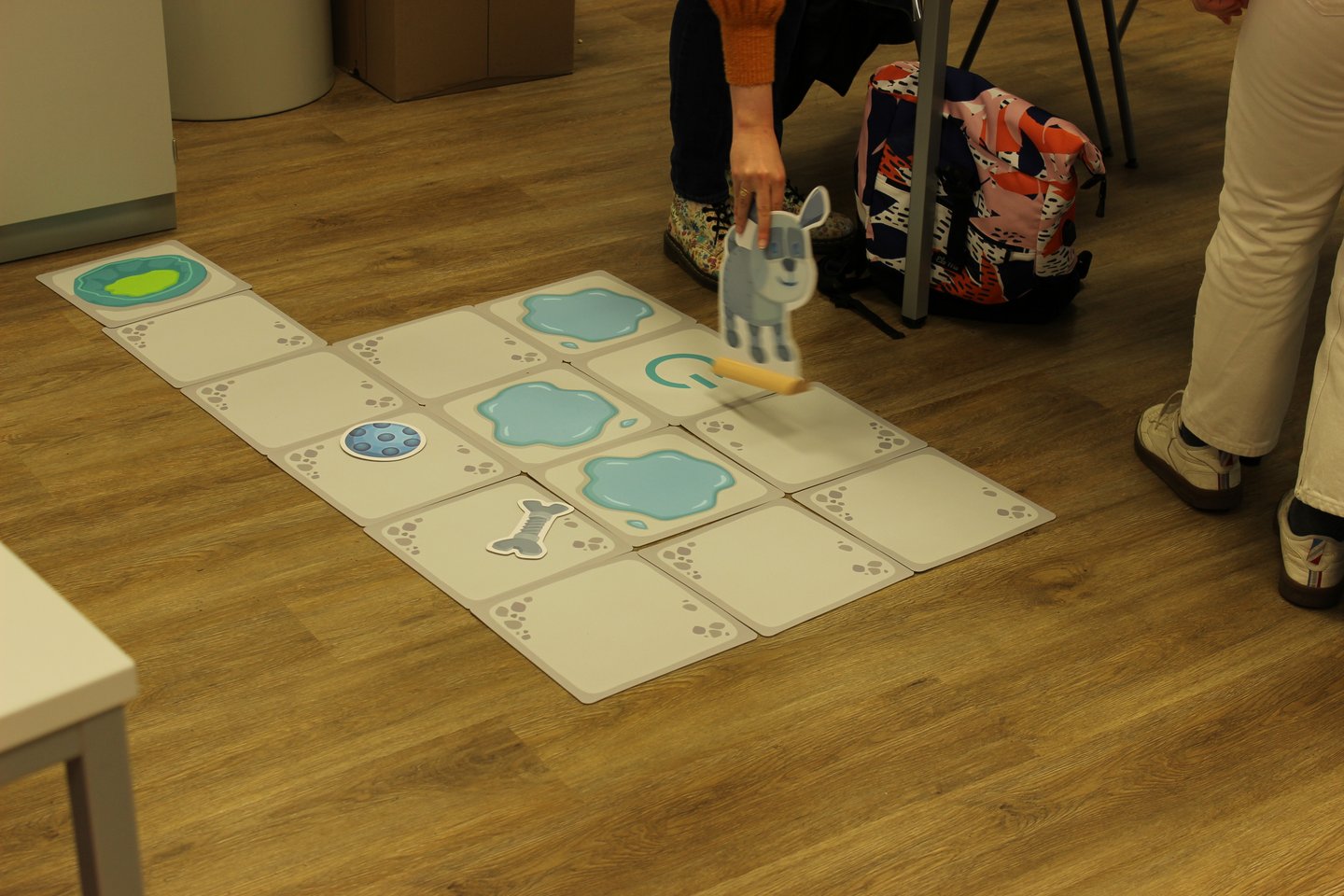 Es handelt sich um ein Lernspiel auf dem Fußboden. Große Karten auf dem Boden stellen Pfützen, Startposition, Hundekorb und normale Schrittflächen da. Auf einer Karte liegt ein Knochen, auf einer ein Ball. Eine Person bewegt eine große Hundefigur entlang der Karten.